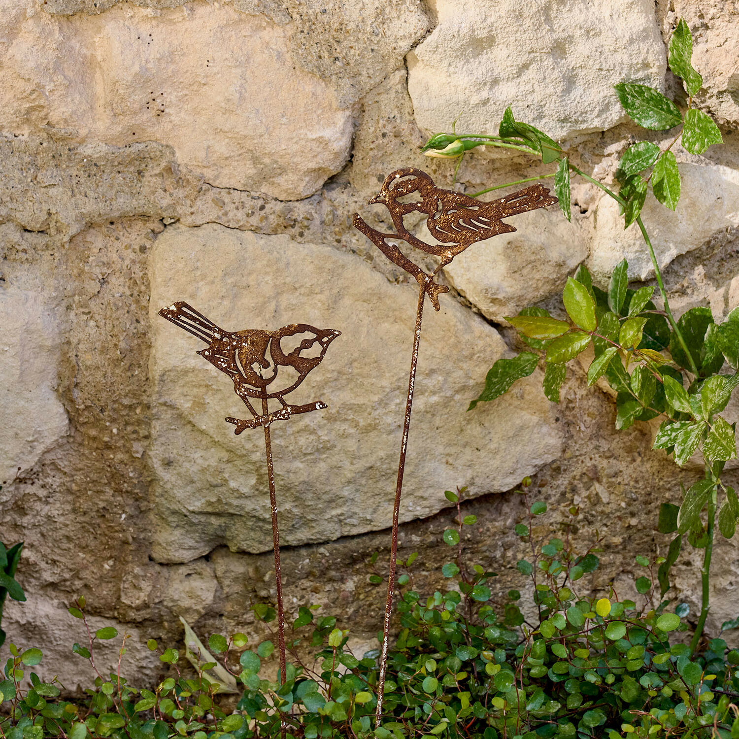 Décorations de jardin en métal rouillé en forme d'oiseaux sur tiges devant un mur en pierre