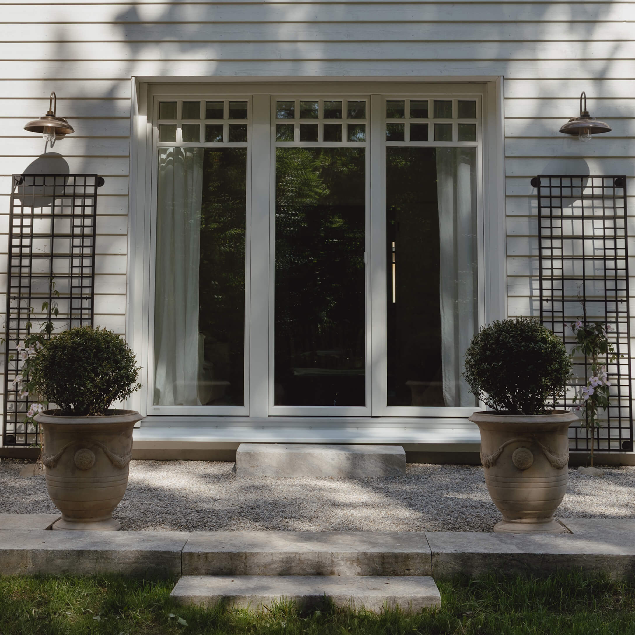 Entrée de maison avec porte blanche et deux grandes jardinières en pierre avec plantes vertes sur gravier