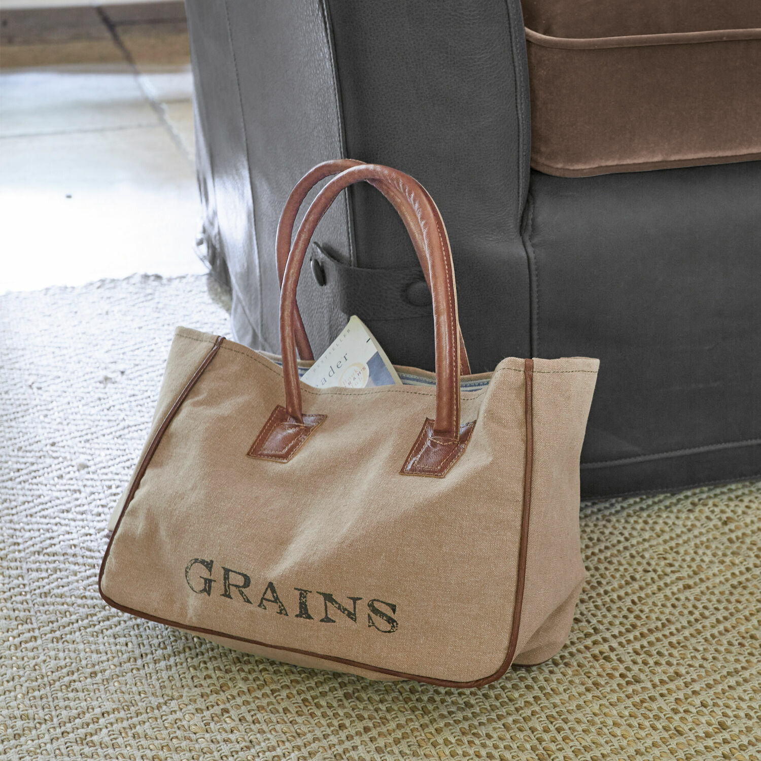 Sac Grains Sac beige en tissu avec poignées en cuir marron et inscription GRAINS posé sur un tapis près d'un canapé