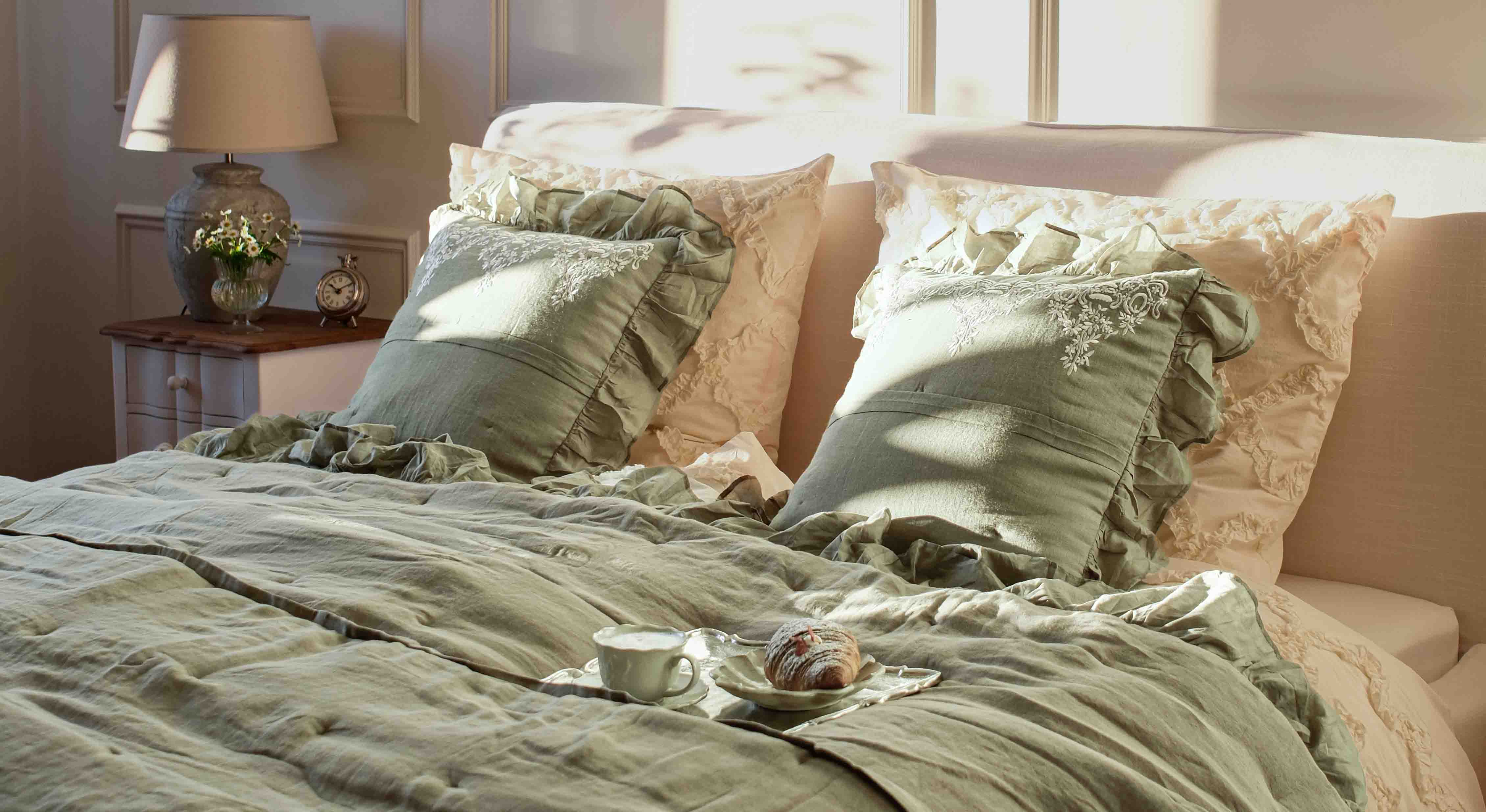 Linge de lit vert en lin avec coussins décoratifs et taies crème dans une chambre avec lampe et horloge