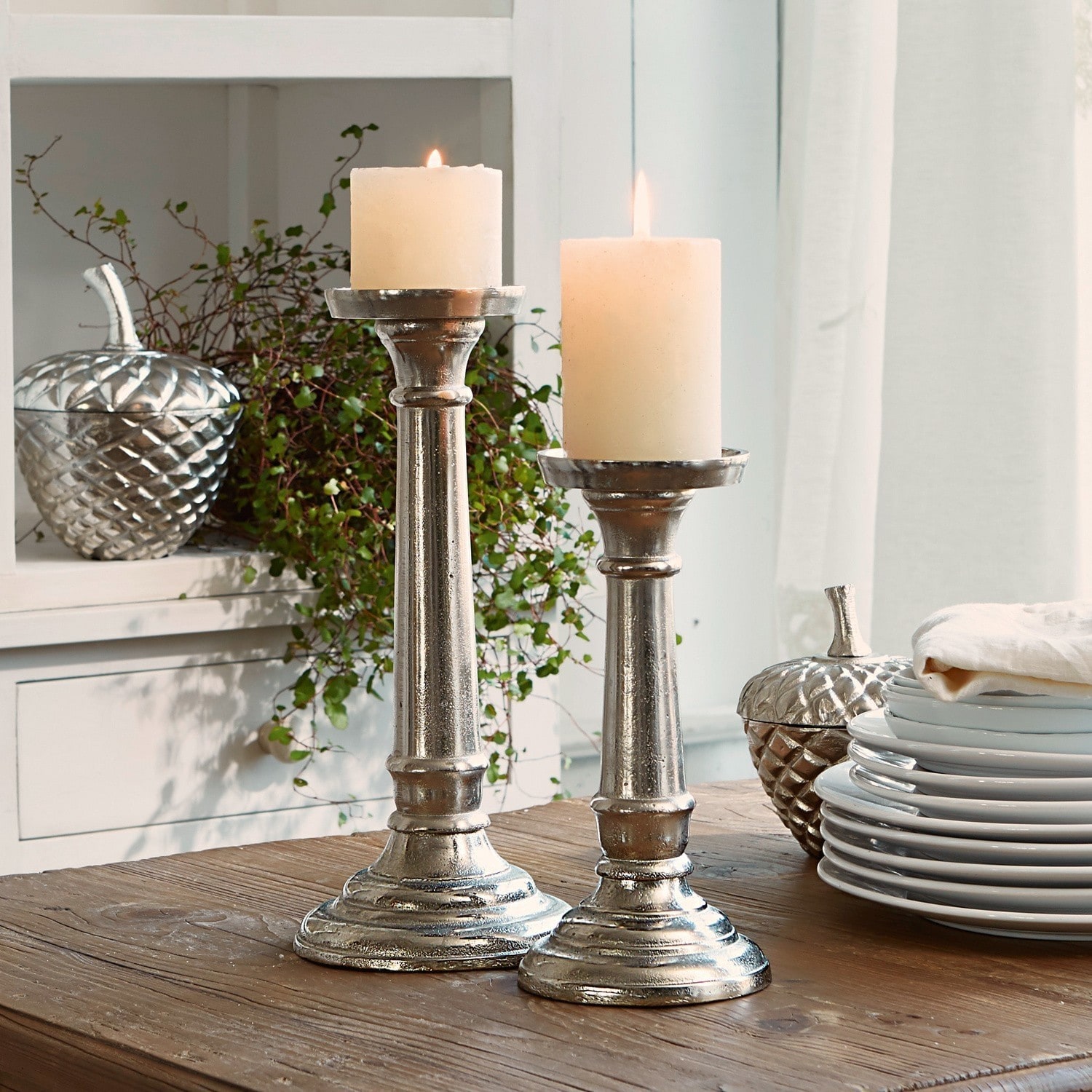 Lot de 2 chandeliers Symone Deux bougeoirs en métal argenté avec bougies blanches sur une table en bois pour décoration intérieure