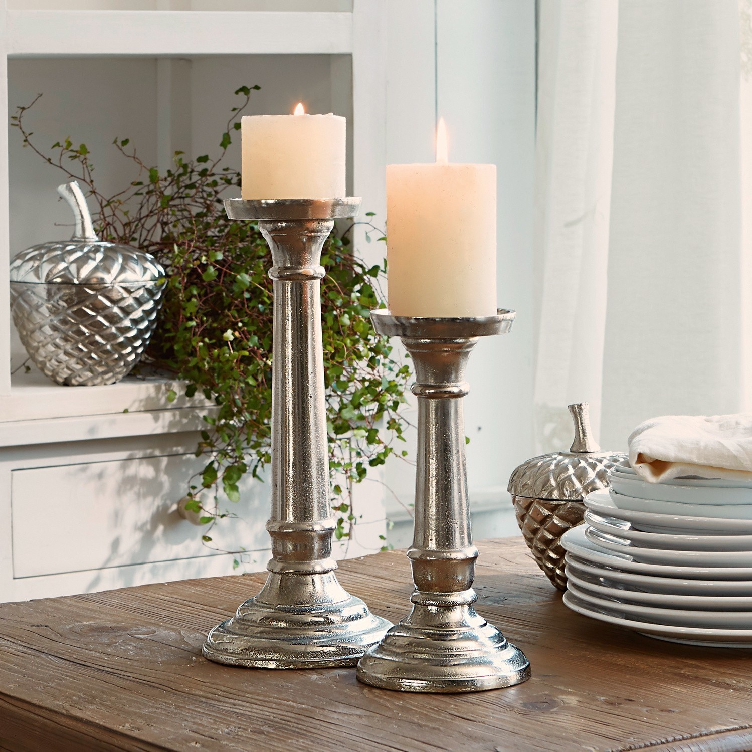 Lot de 2 chandeliers Symone Deux bougeoirs en métal argenté avec bougies blanches sur une table en bois pour décoration intérieure