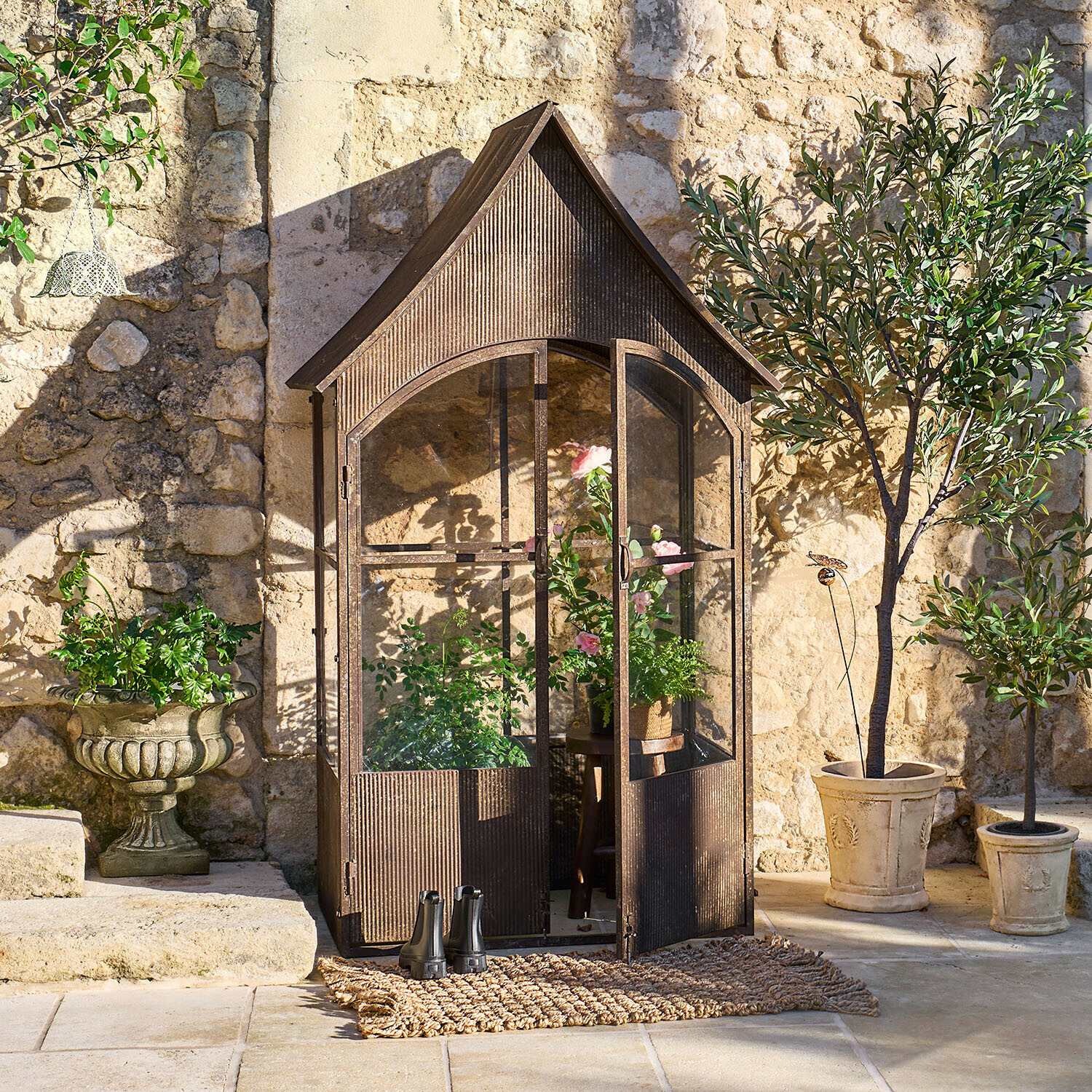 Serre de jardin en métal brun style vintage avec plantes et pots devant un mur en pierre ensoleillé