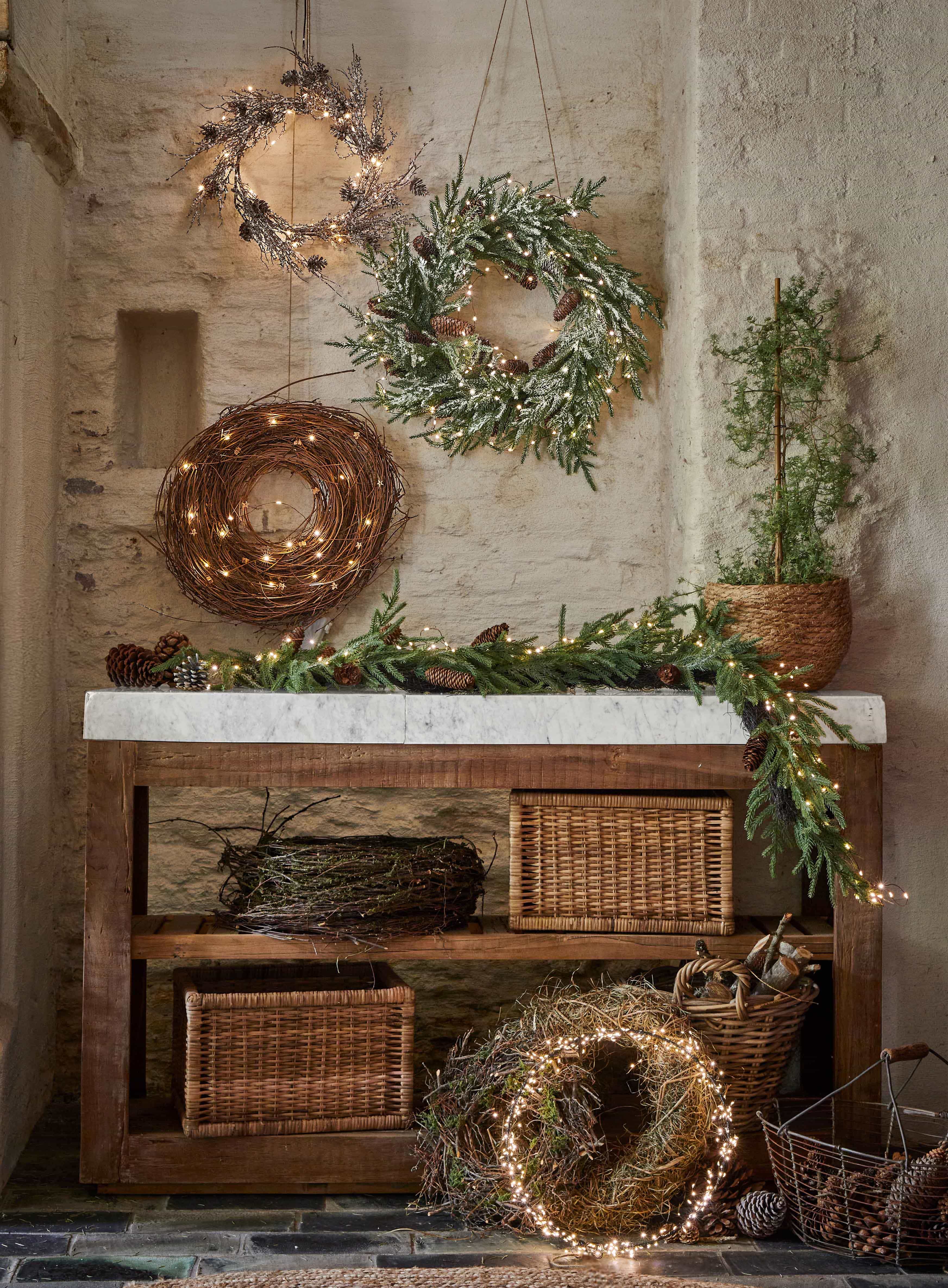 Couronnes décoratives de Noël en branches, pommes de pin et lumières sur une table en bois dans un intérieur rustique