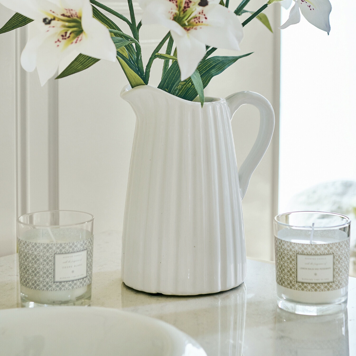 Pichet Belair Pichet en céramique blanche cannelée avec fleurs de lys et deux bougies sur une table en marbre