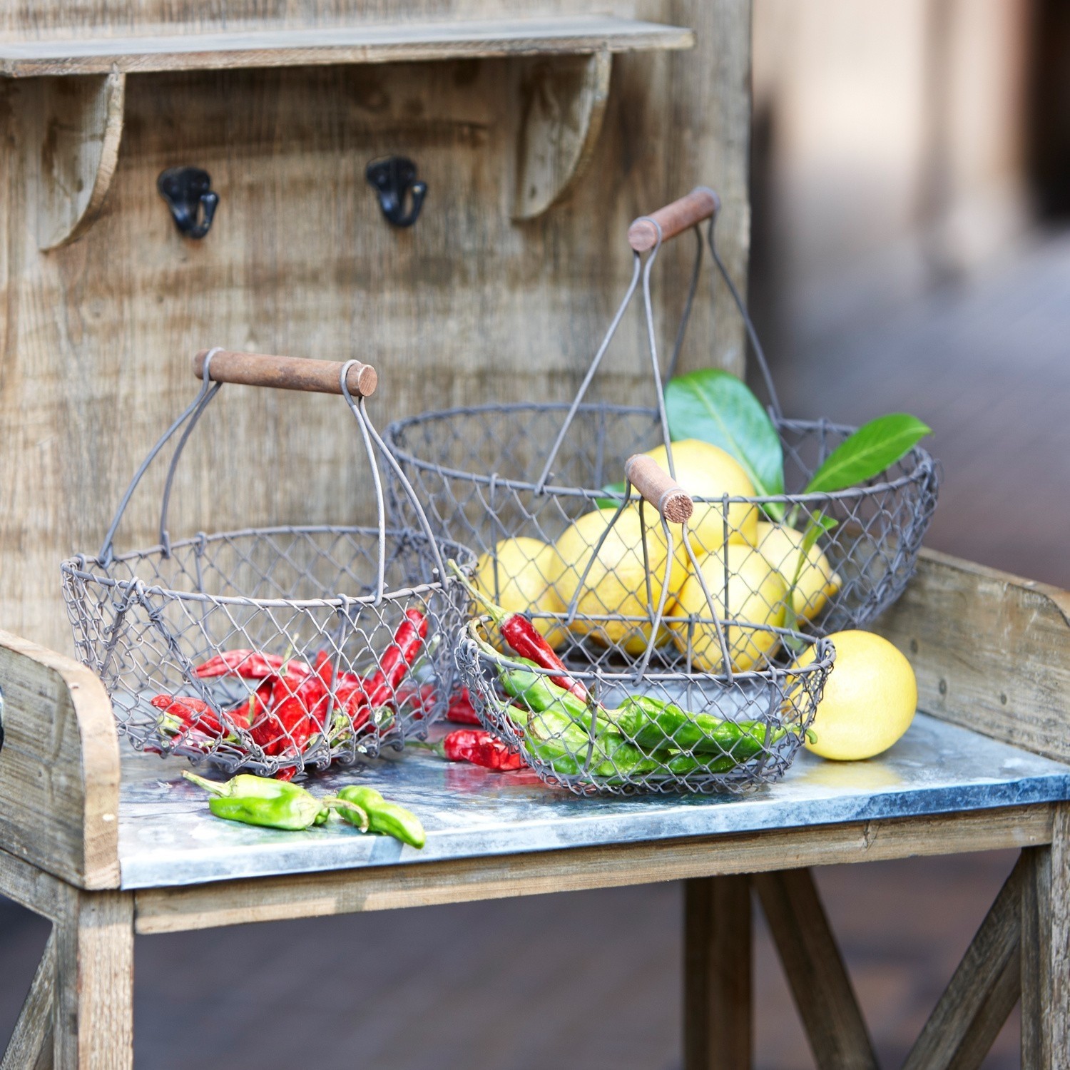 Lot de 3 paniers en grillage Baker Trois paniers métalliques avec poignées en bois contenant des piments rouges, verts et des citrons sur une table en bois