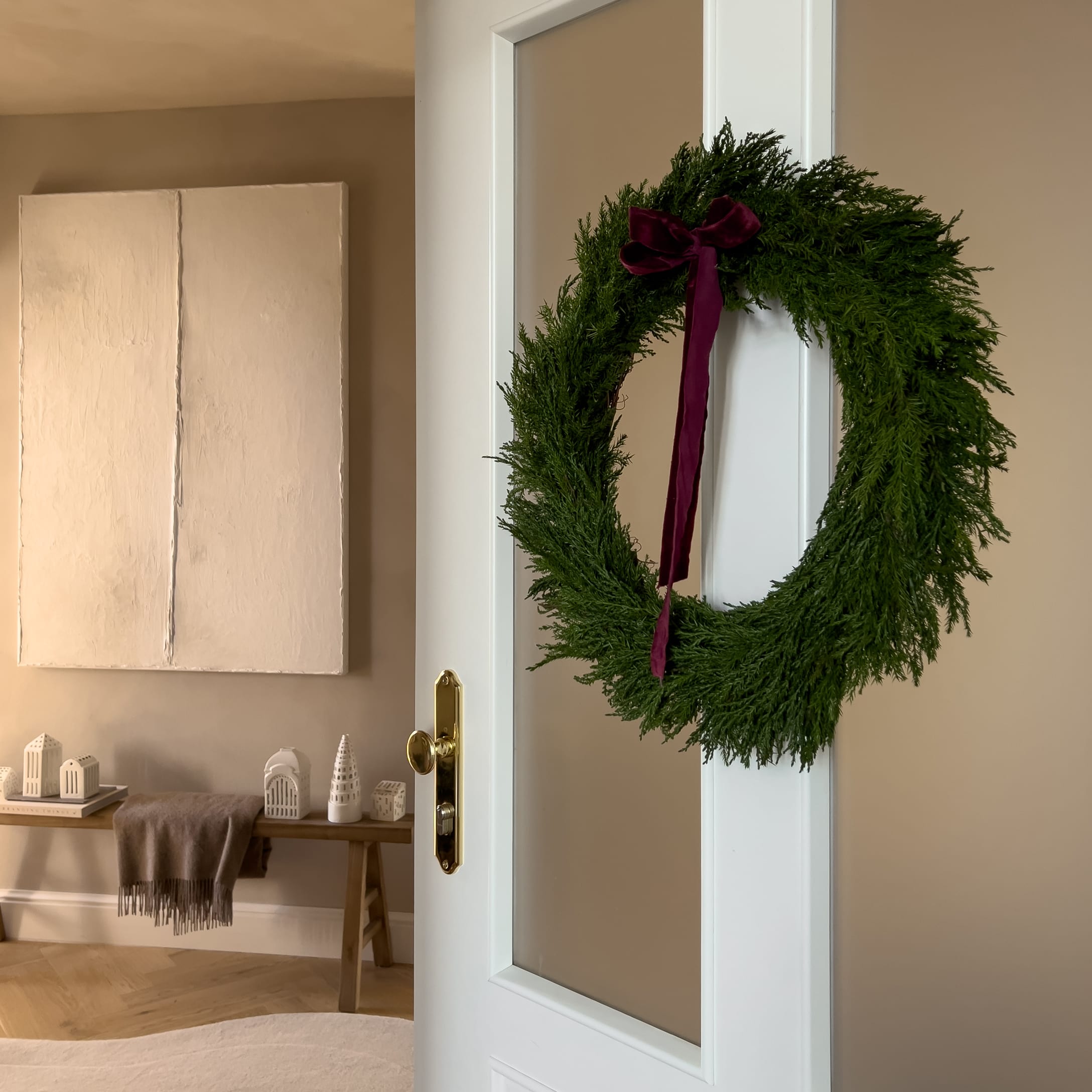 Couronne de Noël verte avec un ruban bordeaux accrochée à une porte blanche dans un intérieur chaleureux
