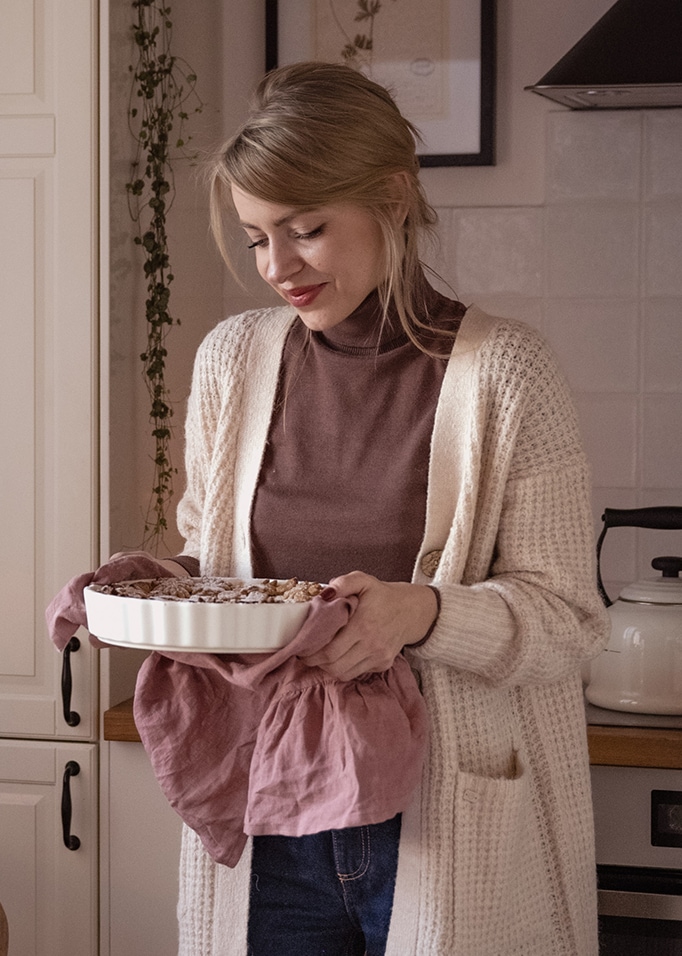Femme en cardigan beige tenant un plat à four en céramique blanche avec un torchon rose dans une cuisine