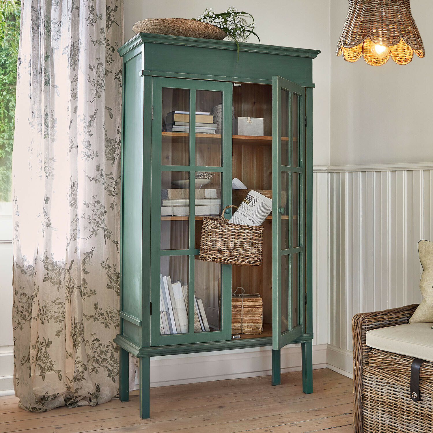 Armoire Faymere Vitrine en bois vert avec portes vitrées et panier en osier dans un salon lumineux au style naturel