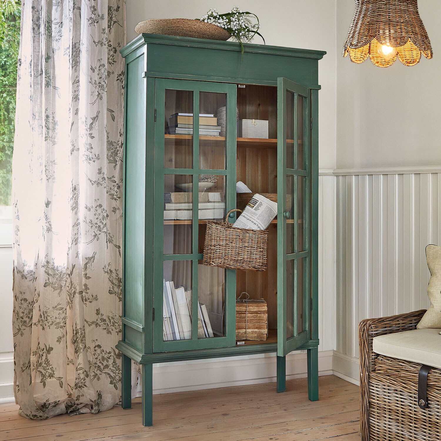 Armoire Faymere Vitrine en bois vert avec portes vitrées et panier en osier dans un salon lumineux au style naturel