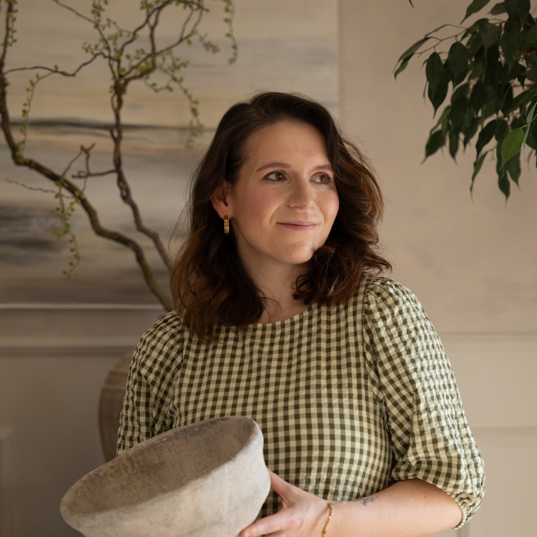 Femme en robe à carreaux verts tenant un grand bol en céramique grise dans un intérieur lumineux avec des plantes