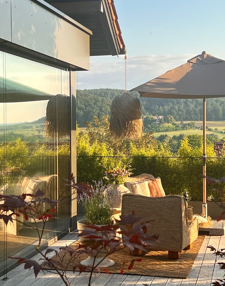 Terrasse confortable avec canapé beige, parasol et plantes devant un paysage verdoyant et une forêt.