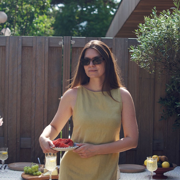 Femme en robe jaune en lin tenant une assiette de pastèque devant une clôture en bois et une table avec des fruits