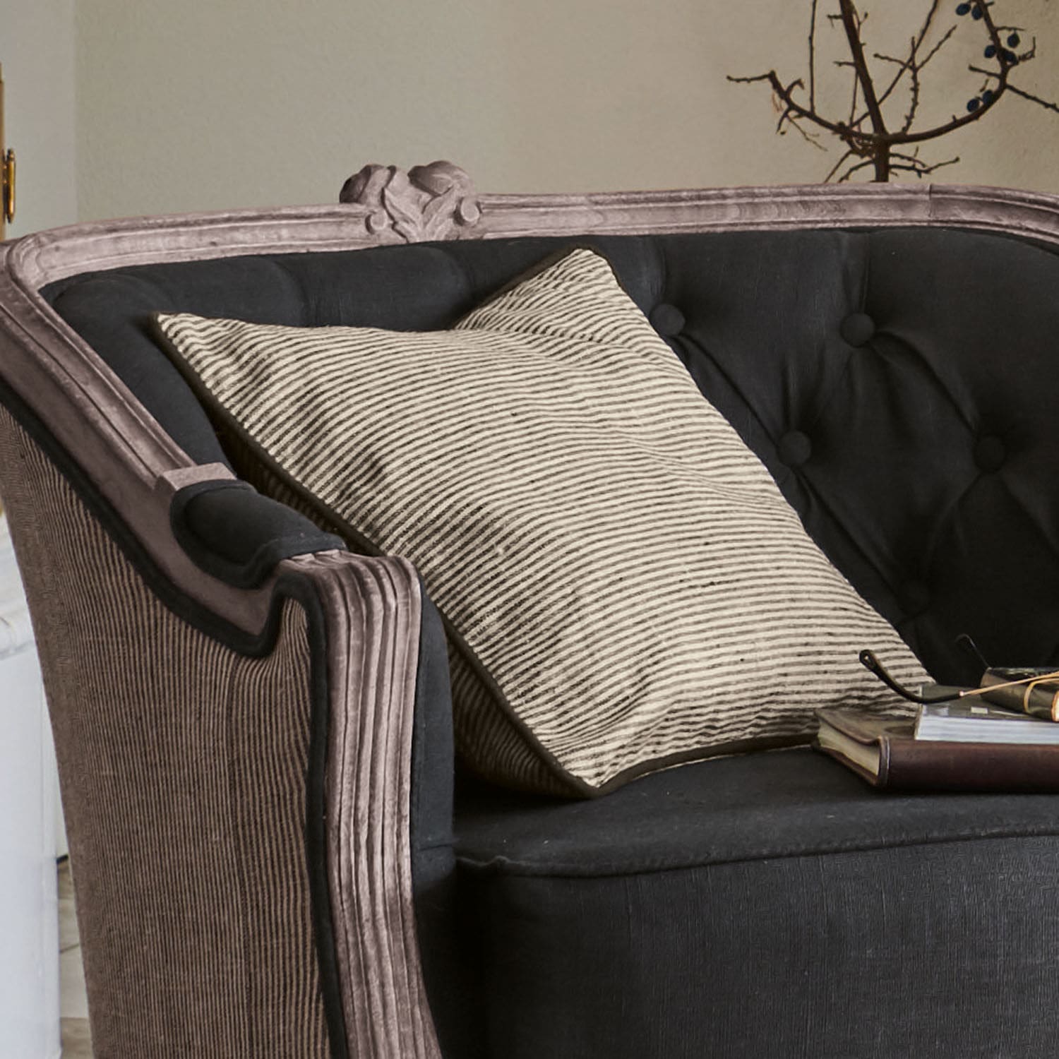 Housse de coussin Aspet Canapé noir capitonné avec coussin beige rayé et livre posé, ambiance vintage élégante