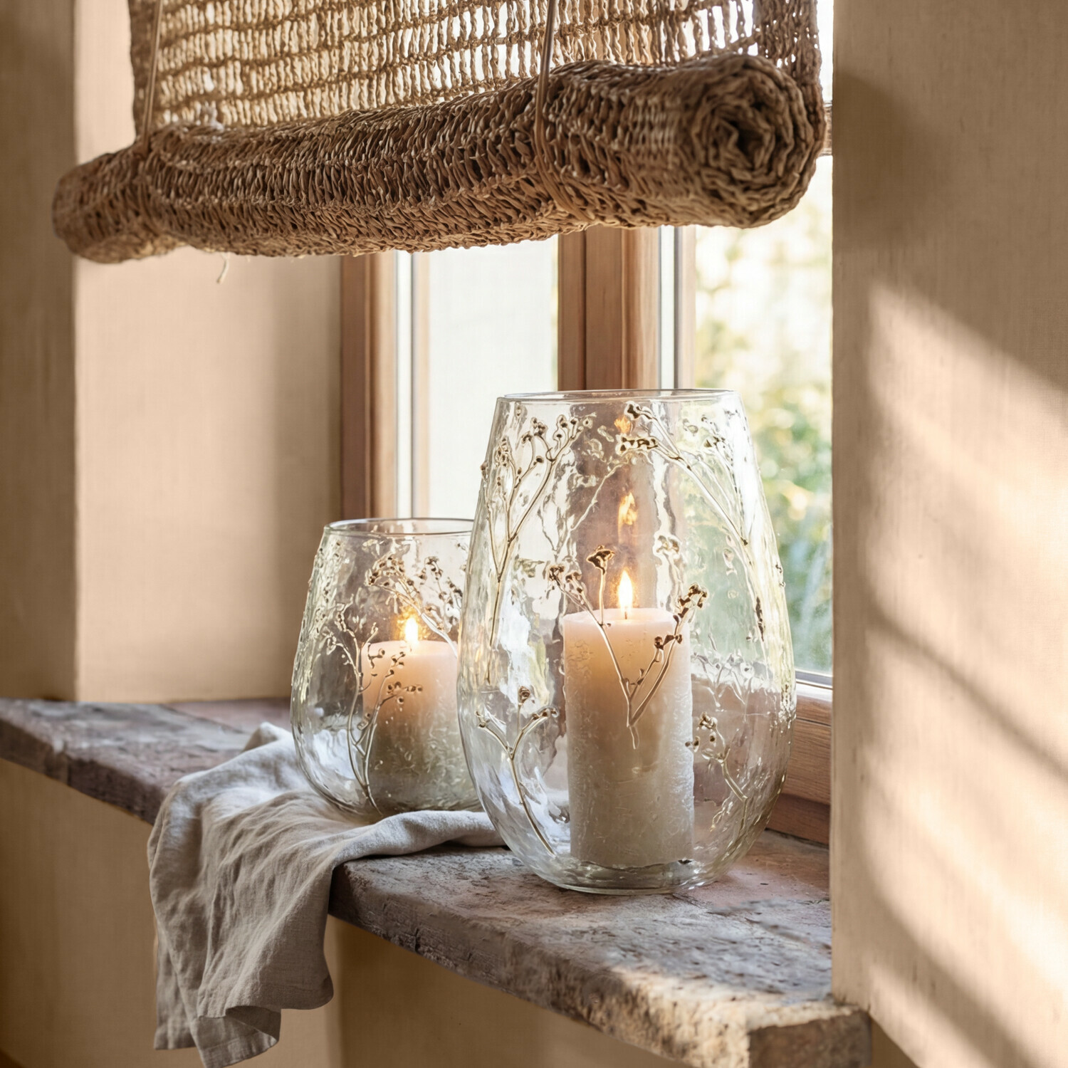 Lot de 2 photophores Elorisse Photophores en verre avec motifs floraux en relief et bougies blanches sur un rebord en pierre au soleil