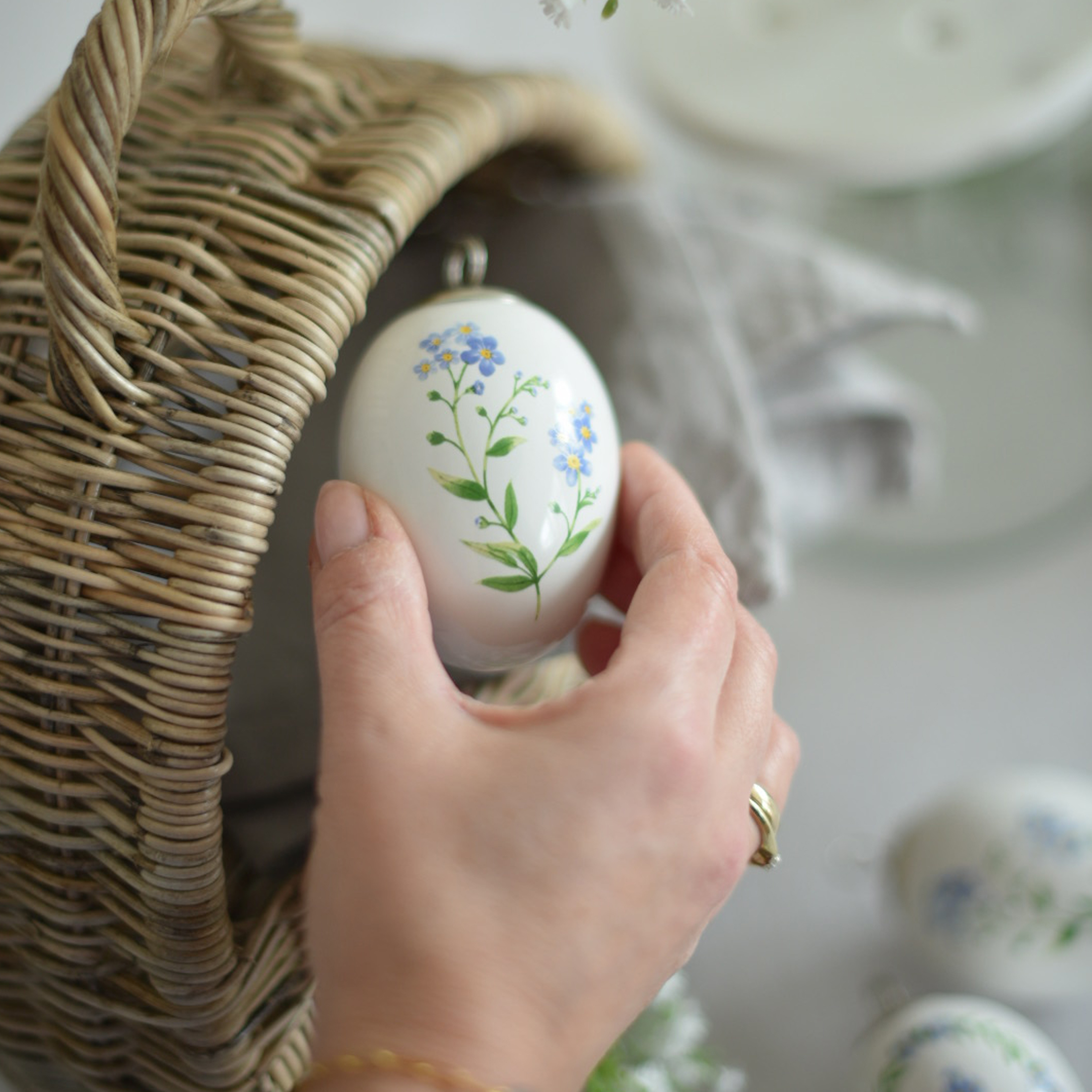 Main tenant une décoration de Pâques en céramique blanche avec fleurs bleues près d'un panier en osier