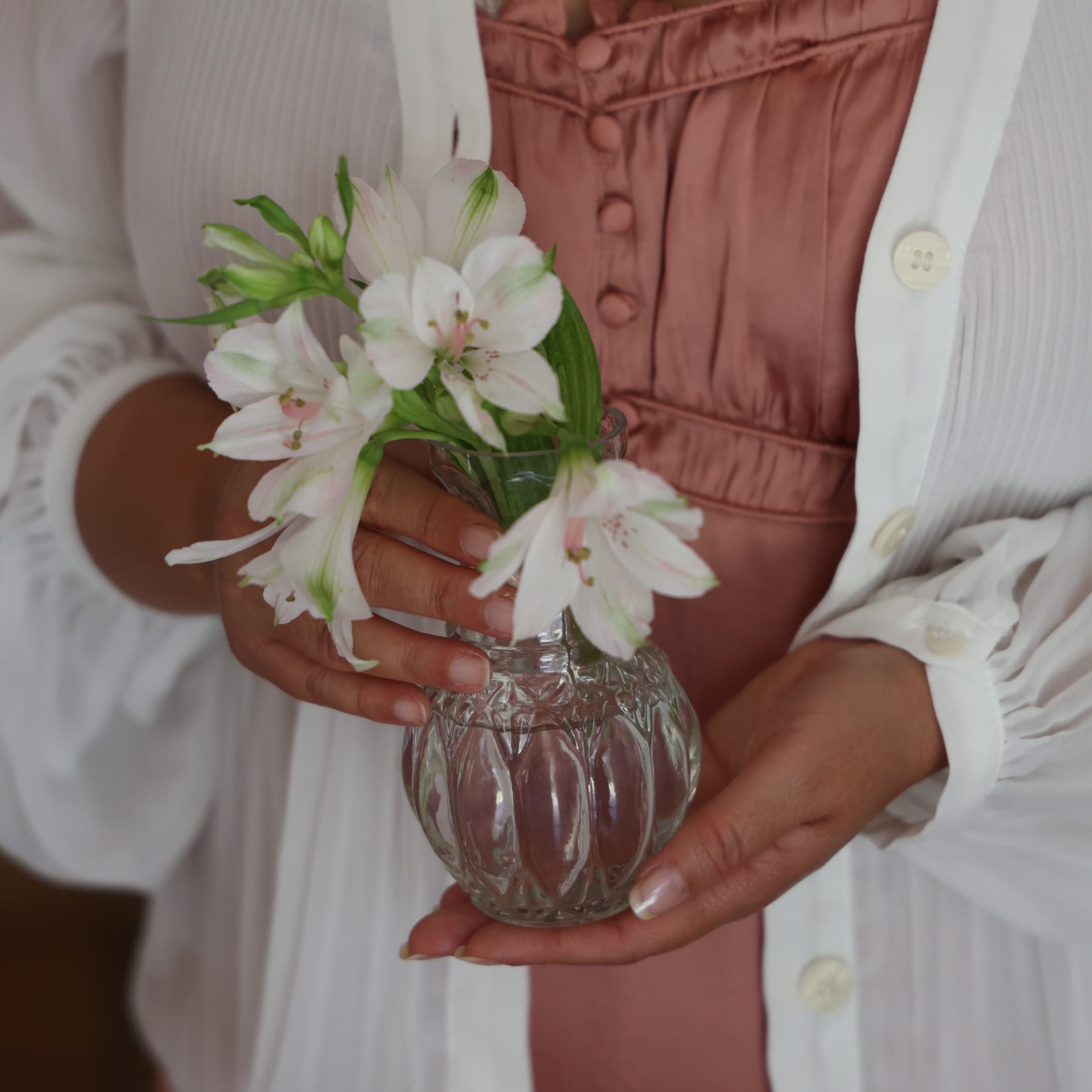 Femme tenant un vase en verre transparent avec des fleurs blanches devant une robe rose et un gilet blanc