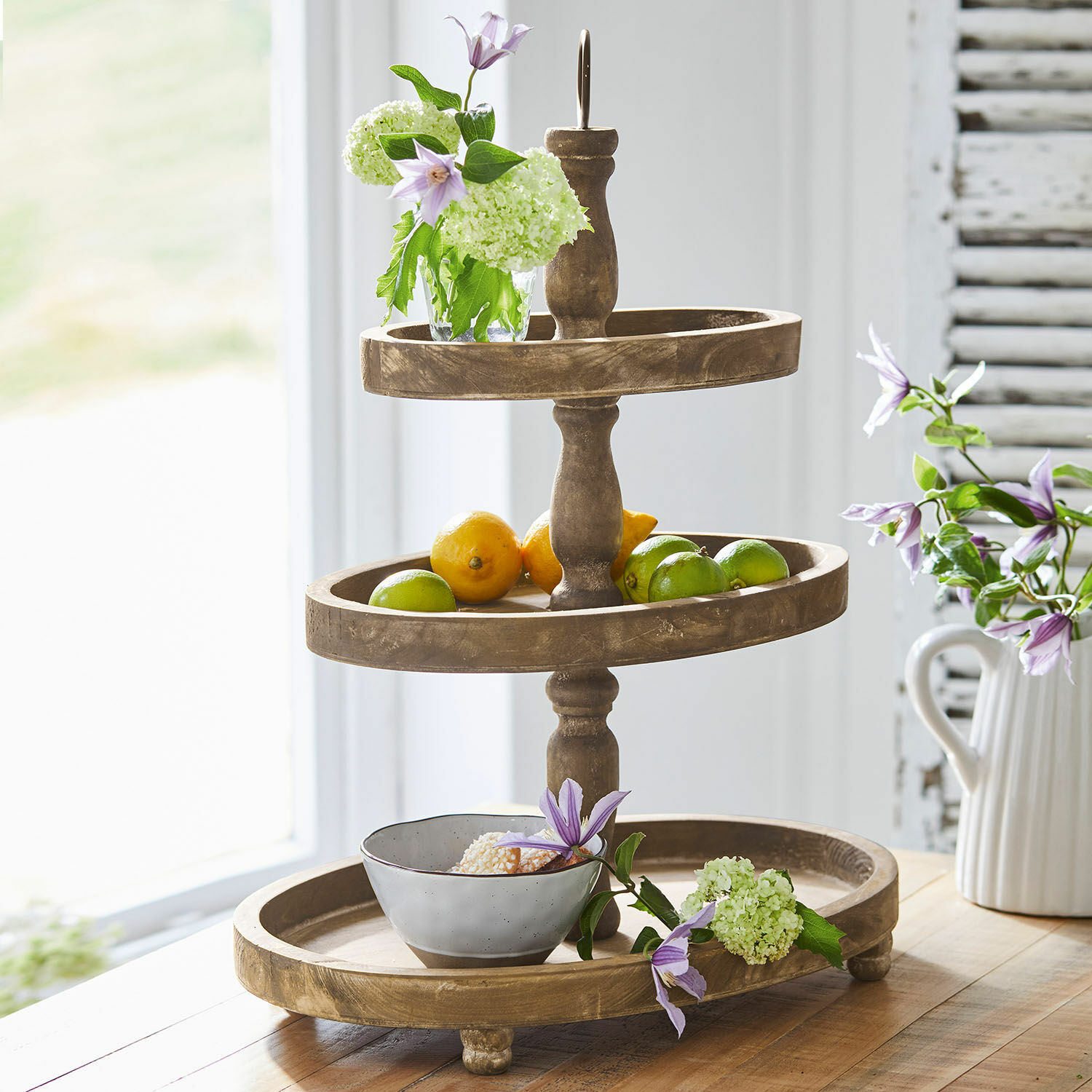 Présentoir à étages Olathe Présentoir à trois niveaux en bois avec fruits et fleurs sur une table en bois dans une pièce lumineuse
