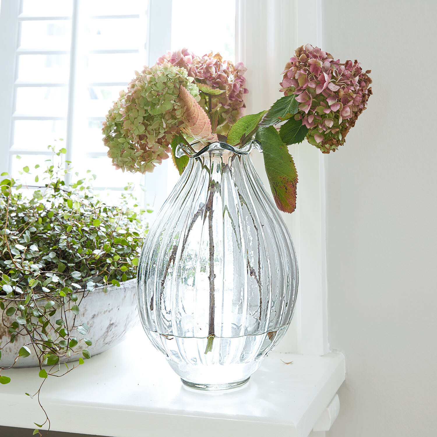 Vase Jardin Lorrain Vase en verre transparent cannelé avec hortensias roses et verts posé sur un rebord de fenêtre blanc
