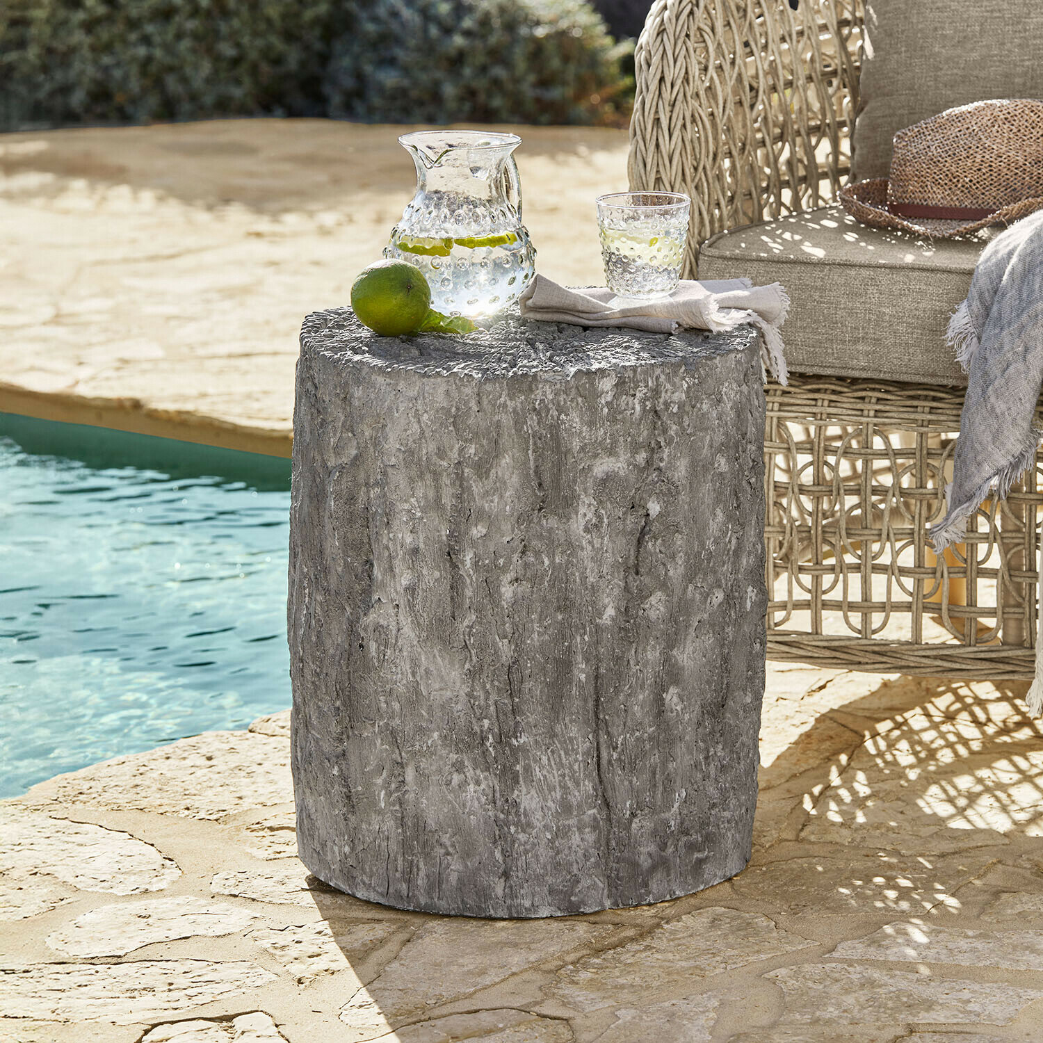 Table d'appoint grise en béton à texture bois près d'un fauteuil en osier au bord de la piscine