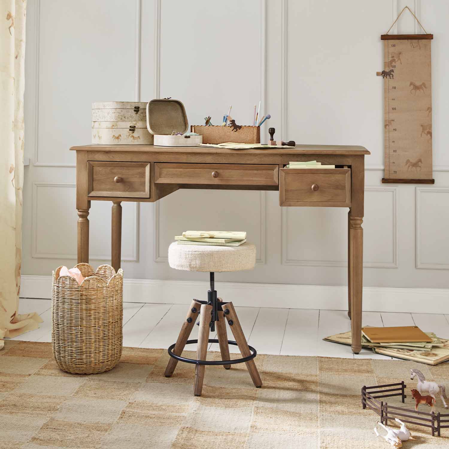Bureau en bois avec trois tiroirs et tabouret beige devant un mur clair décoré de motifs chevaux